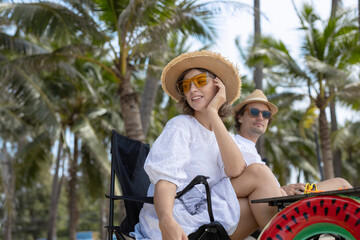 Happy couple enjoying tropical summer vacation at the beach. Smiling woman in white dress and straw hat relaxing in wheelchair with palm trees background on sunny day.