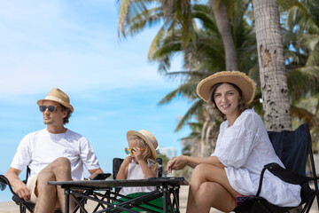 Happy family enjoying summer beach vacation together. Parents and child wearing hats and sunglasses sitting on chairs under palm trees, relaxing and smiling on a bright sunny day.