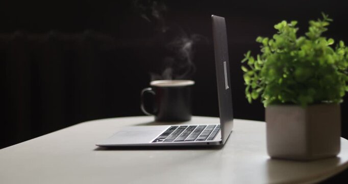 Close-up of laptop on table with hand in red sweater, taking a black  coffee cup onto table surface abd typing on keyboard, blurred background
