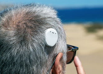 A man with grey hair wearing glasses and using a hearing aid. The device is visible behind the ear,...