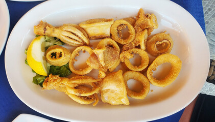 A plate of deep-fried squid rings. The golden rings and pieces are coated in a crispy breading. The dish is garnished with a lemon wedge and green leaf lettuce.