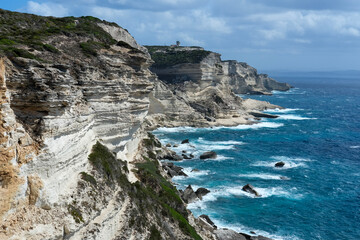 Sheer stratified cliffs above a choppy blue sea with a lighthouse on the distant point.