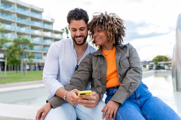 Diverse couple smiling, looking at smartphone together outdoors