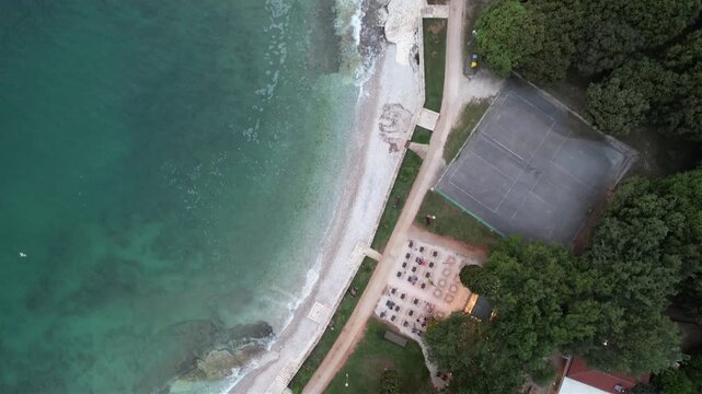 Aerial drone view of a rocky sunset beach coastline in Lanterna peninsula, Istria, Croatia, with crystal clear turquoise water, surf waves and lush green forest above the cliffs.