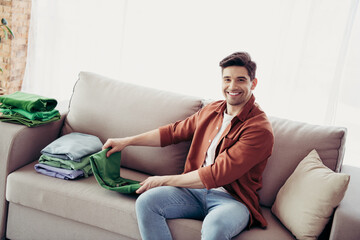 Fototapeta premium Young man folding laundry on sofa at home, smiling during household chores in the living room, enjoying domestic tasks