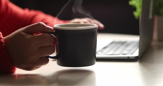 Close-up of laptop on table with hand in red sweater, taking a black  coffee cup onto table surface abd typing on keyboard, blurred background
