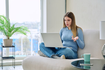 Young woman looking at laptop screen talking having video call sitting on chair at home