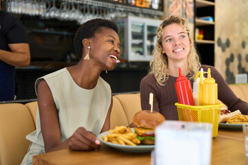 Diverse women friends laughing eating hamburgers at restaurant