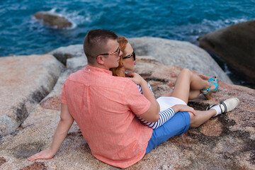 A young couple sits closely on sun-warmed granite rocks by the ocean; the man in an orange shirt holds the woman, who wears a striped top, white shorts, and sunglasses, looking up at him with a loving