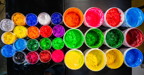 A collection of plastisol inks stored in sturdy barrels and glass jars, each filled with rich and vibrant colors red, blue, yellow, green, pink, orange, purple, and white in screen printing factory