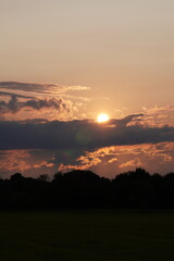Scenic sunset with glowing sun above dark treeline and dramatic evening clouds, peaceful natural landscape with warm colors.