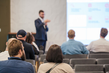 Presenter in suit delivers talk to attentive audience in conference room with projection screen showing slides