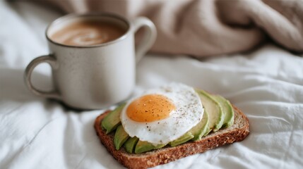 A well-prepared breakfast features a slice of whole grain bread topped with sliced avocado and a sunny side up egg accompanied by a warm cup of coffee on a cozy bed.