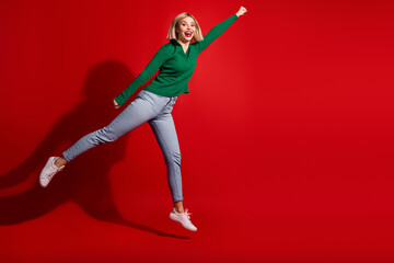 Energetic young woman jumping joyfully in green sweater and jeans on red background with positive expression and gesture © deagreez