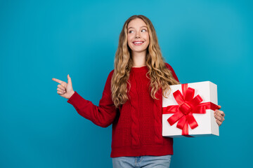 Fototapeta premium Adorable young woman in a red sweater holding a gift box wrapped with a red bow against a vibrant blue background