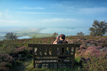A woman on a bench looks through binoculars in front of a picturesque lake. Vane Hill, Loch Leven, Scotland