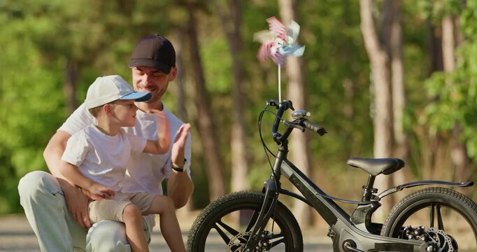 Caring dad hugs son tightly beside bicycle during joyful pause between rides. Boy leans against loving parent with smile while father holds embrace warmly in park