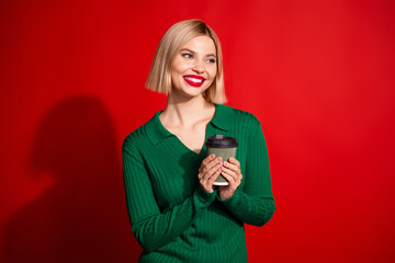 Naklejka premium Lovely stylish blonde woman in a green sweater holding a coffee cup against a bright red background, smiling happily