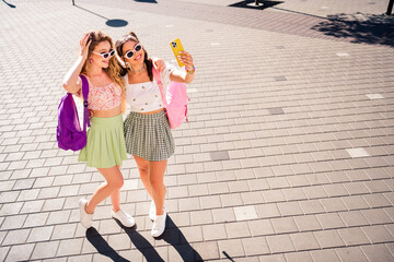 Two stylish young friends take a sunny selfie on a city street with backpacks and sunglasses capturing a fun casual travel moment