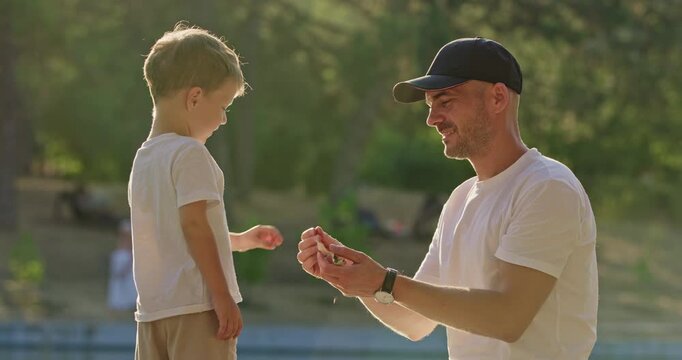 Caring young father gives yellow leaf to son while standing on park path. Preschooler child takes leaf throwing item into water preparing to hold another piece