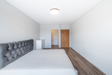 Home Bedroom Interior. King Size Double Bed, Brown Wooden Exit Door. White Walls and Ceiling. LED Liaght. Dark Hardwood Floor. White Chest of Drawers