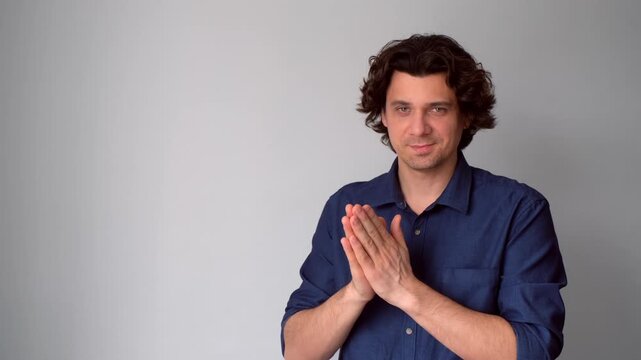 Sneaky scheming curly man rubbing hands plotting cunning idea on gray background looking at camera. Middle aged brunet guy smirking and conspiring villain plan with greedy sly face expression.