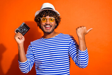 Cheerful young man in a striped shirt holding a vintage camera and pointing on an orange background