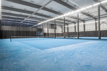 Modern Indoor Tennis Court With Clean Blue Flooring and Ample Lighting. Indoor tennis court featuring bright blue flooring, net setup, and illuminated by large lights in a spacious sports facility.