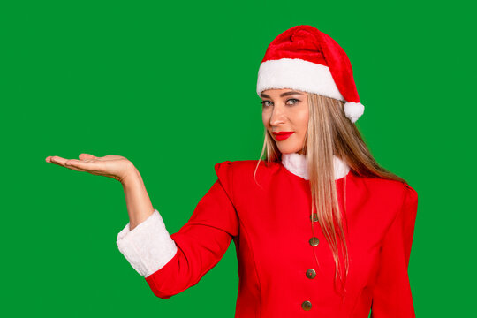 A woman in a Mrs. Claus costume is presenting an object with her hand, standing in front of a green screen backdrop