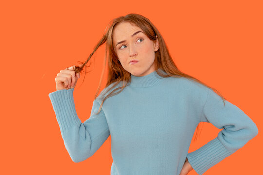 Pensive redhead twists a strand of her hair against an orange background, contemplating something