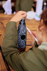 Child decorating a reusable water bottle with a white marker during an outdoor art workshop or creative activity