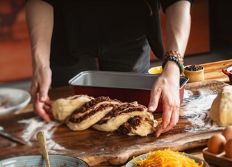 A person carefully transfers a beautifully braided Cozonac dough onto a baking sheet, ready for baking. The scene is set on a rustic wooden board with flour dusting the surface