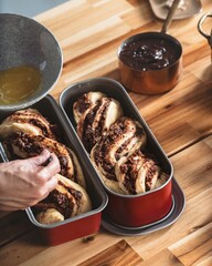 A person is carefully arranging the risen Cozonac dough in the baking pans before putting it in the oven. There is also a bowl of egg wash and a pot of chocolate nearby