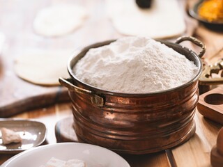 A rustic scene showing a copper pot overflowing with flour, surrounded by dough, yeast, and other baking necessities on a wooden table, preparing for homemade baking