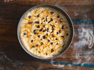 A freshly baked, decadent cozonac rests in its baking pan. Orange zest and chunks of dark chocolate are scattered across its creamy surface, set against a rustic wooden backdrop