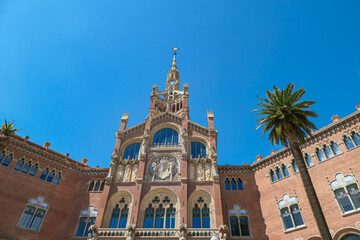 Hospital de Sant Pau’s impressive main building stands under vivid blue sky, palm trees framing its elaborate modernist design, highlighting Barcelona’s renowned architectural legacy.