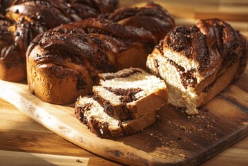 Freshly baked Cozonac bread sits on a wooden board, partially sliced to reveal chocolate swirls. Sunlight enhances its texture. A delightful homemade treat ready to eat
