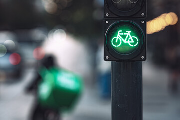 Close-up of green bicycle traffic light signal illuminated with food delivery cyclist riding on urban street in background