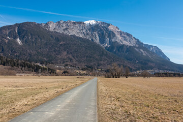A straight paved road leads through open fields toward the imposing Dobratsch mountain, snow-capped and rising above Nötsch im Gailtal, Carinthia, Austria, under a clear blue sky in the Gailtal Alps.