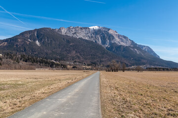Long straight road leads across open meadows toward the imposing Dobratsch mountain in Nötsch im Gailtal, Carinthia, Austria, beneath a bright blue sky and surrounded by peaceful rural scenery.