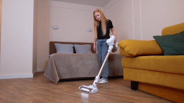 Young teen girl moving cordless vacuum cleaner on wooden bedroom floor, cleaning near yellow sofa and unmade bed, doing home maintenance routines