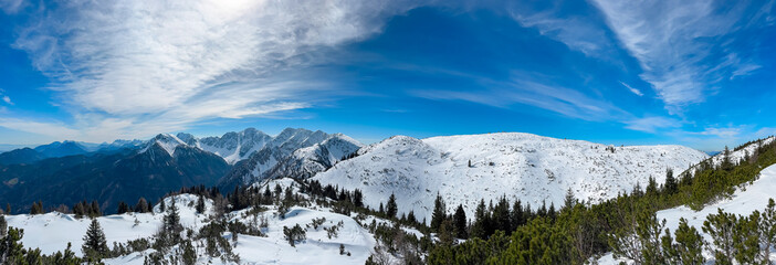 Snow-covered mountains and pine forests stretch beneath a vast blue sky, revealing panoramic views of Hochstuhl, Kosiak, and Weinasch from Bärentaler Kotschna in the Karawanks, Carinthia, Austria.