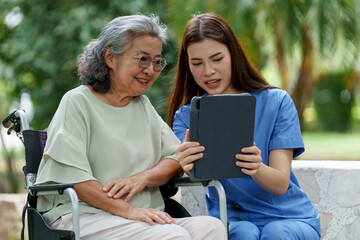 Caring caregiver provides assistance and support to senior in wheelchair while they enjoy moment together, sharing information on tablet in serene outdoor setting