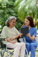 Caregiver provides assistance and support to senior in wheelchair while sharing moment of joy with tablet outdoors. scene reflects compassion and dedication in care