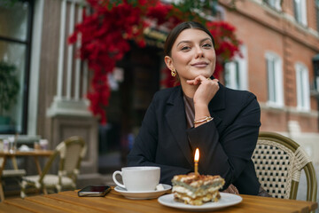 Joyful woman at an outdoor cafe celebrating with a slice of cake featuring a single birthday candle. She wears a smart black blazer and has a cup of tea on the wooden table.