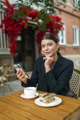 Stylish woman using her smartphone at an outdoor cafe with tea and cake. Dressed in a black blazer and chic outfit, she captures moments in a charming urban setting.