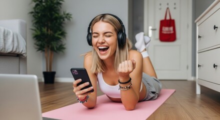 Young woman with headphones laughing while looking at her smartphone at home on a yoga mat
