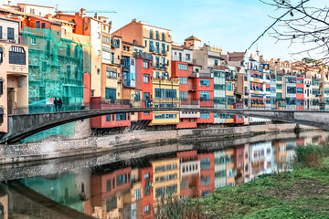 Colorful riverside houses with reflections in the Onyar River and Gomez Bridge, showcasing vibrant Spanish architecture. Girona, Spain - November 28, 2021