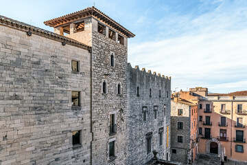 Fototapeta premium Historic Edifici Pia Almoina in Girona, Spain, with medieval stone architecture, crenellated walls and tower, contrasting with colorful houses and blue sky, showcasing rich heritage
