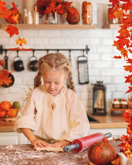 Cute little girl happily rolling out dough with a red rolling pin while baking in a rustic kitchen richly decorated with vibrant orange and red fall leaves. autumn season,  autumn concept,  fall